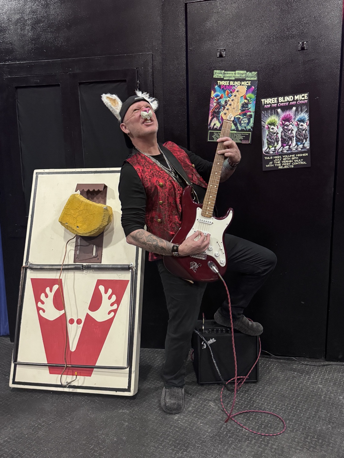 Michael Dargie in mouse ears playing electric guitar beside a giant mousetrap, Three Blind Mice posters behind